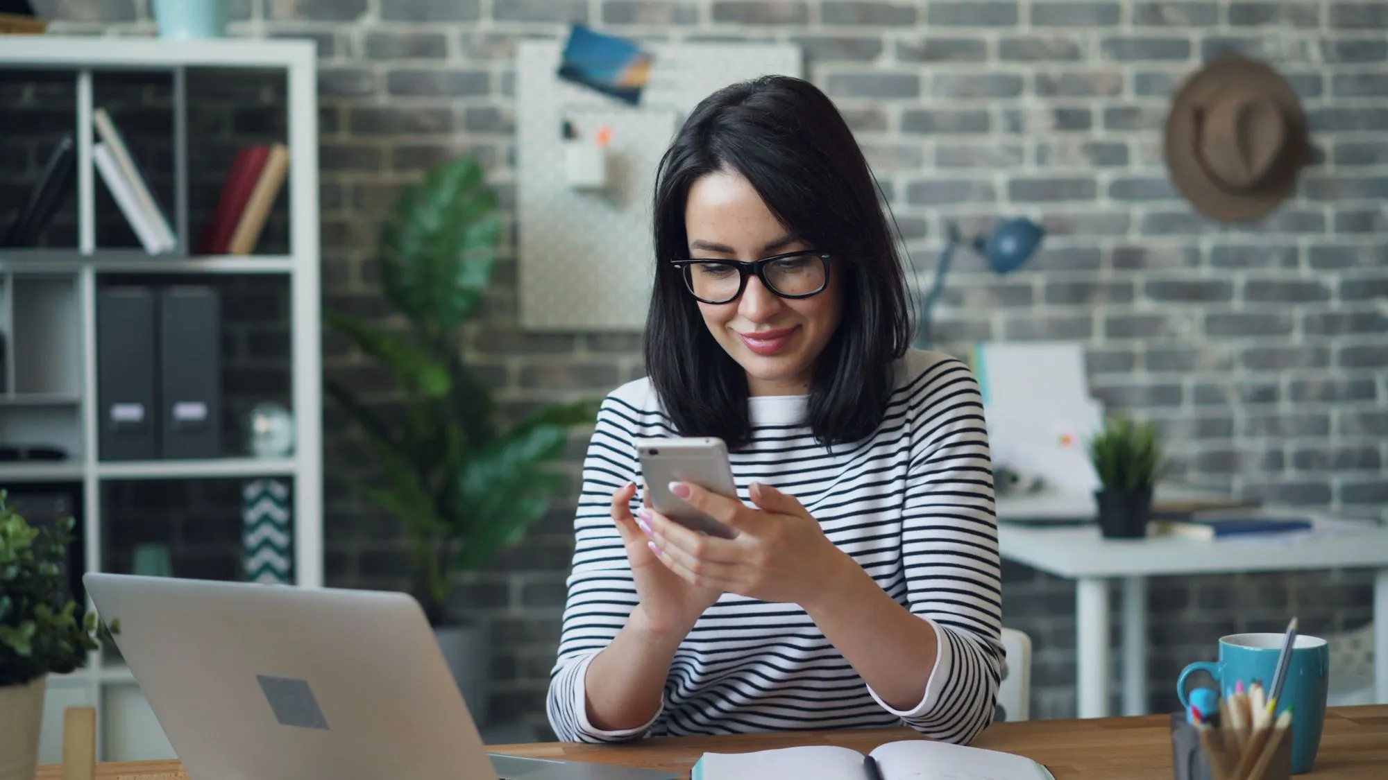 Mulher sentada à mesa de um escritório consulta o celular, com um notebook à frente.